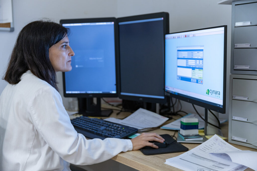 Persona trabajando en una oficina frente a tres monitores, utilizando un teclado y ratón, con documentos y material de oficina sobre el escritorio.